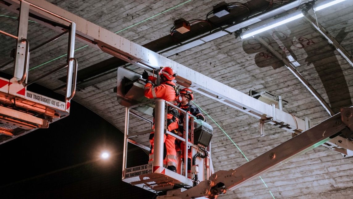 Deux monteurs cablex fixent un signal lumineux LED à une poutre en acier au plafond du tunnel de Honeret.