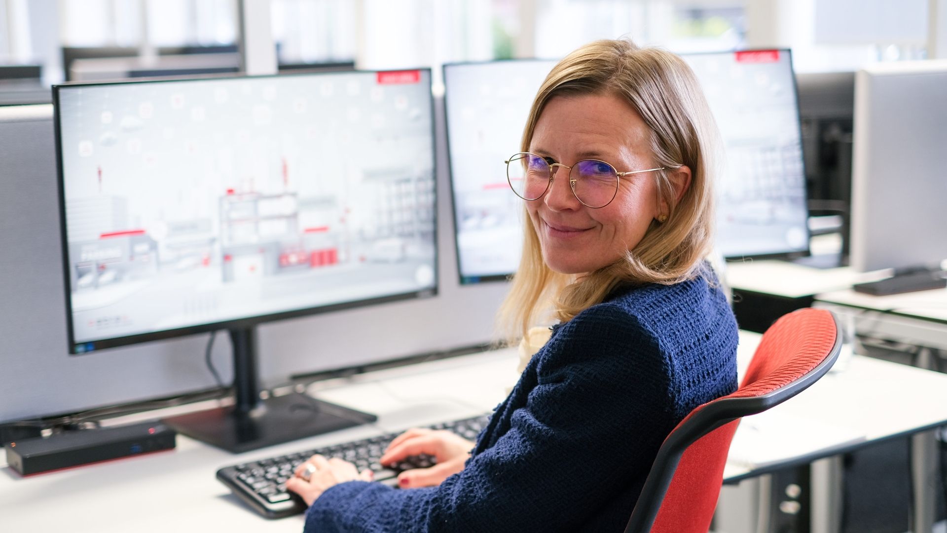 Blonde Frau sitzt auf einem roten Bürostuhl vor dem Computer