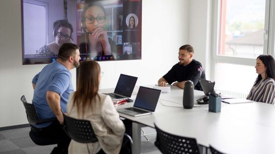 Des collaborateurs de cablex tiennent une réunion hybride dans une salle de réunion moderne. Plusieurs personnes sont connectées par voir numérique sur le grand écran.