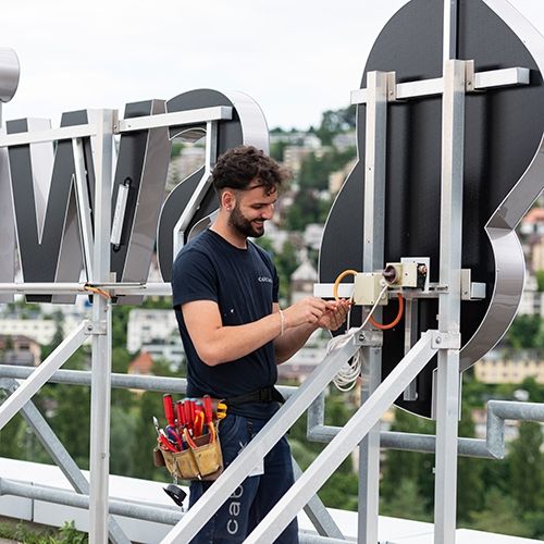 Lernender Elektroinstallateur arbeitet an der Elektroinstallation von Swisscom Logo.